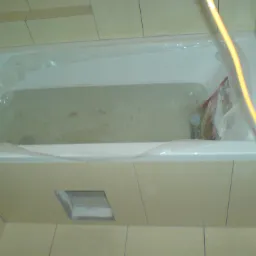 Wanna z obudową w trakcie montażu, zabezpieczona folią, widoczny otw&oacute;r rewizyjny w obudowie, płytki beżowe na ścianach.